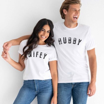 a man and woman laughing, standing affectionately, wearing matching hubby wifey t-shirts in white