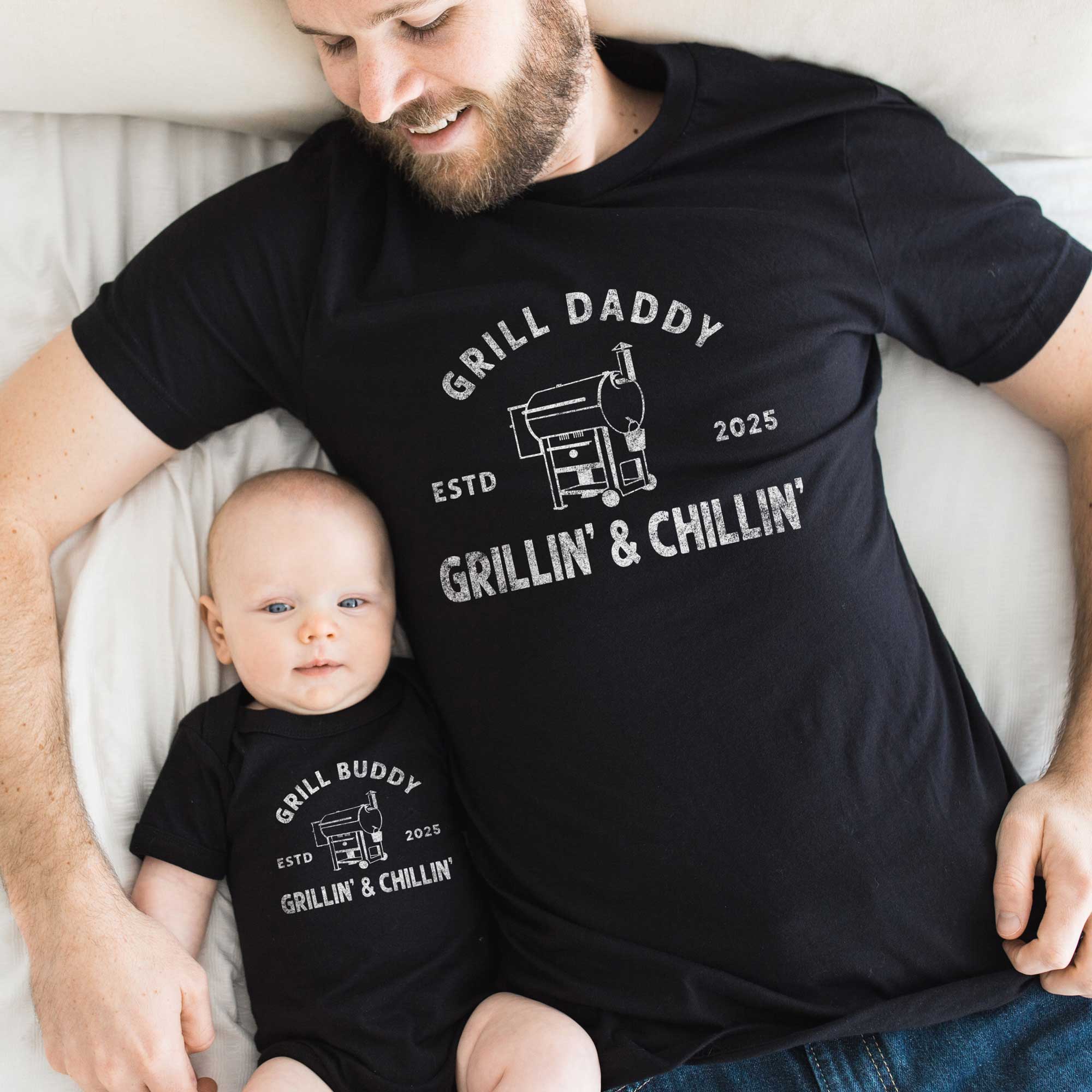 A dad and baby lying together on a bed wearing black tops. The baby is wearing a Grill Buddy Onesie: Smoker Grill, the dad is wearing a Grill Daddy t-shirt: Smoker Grill
