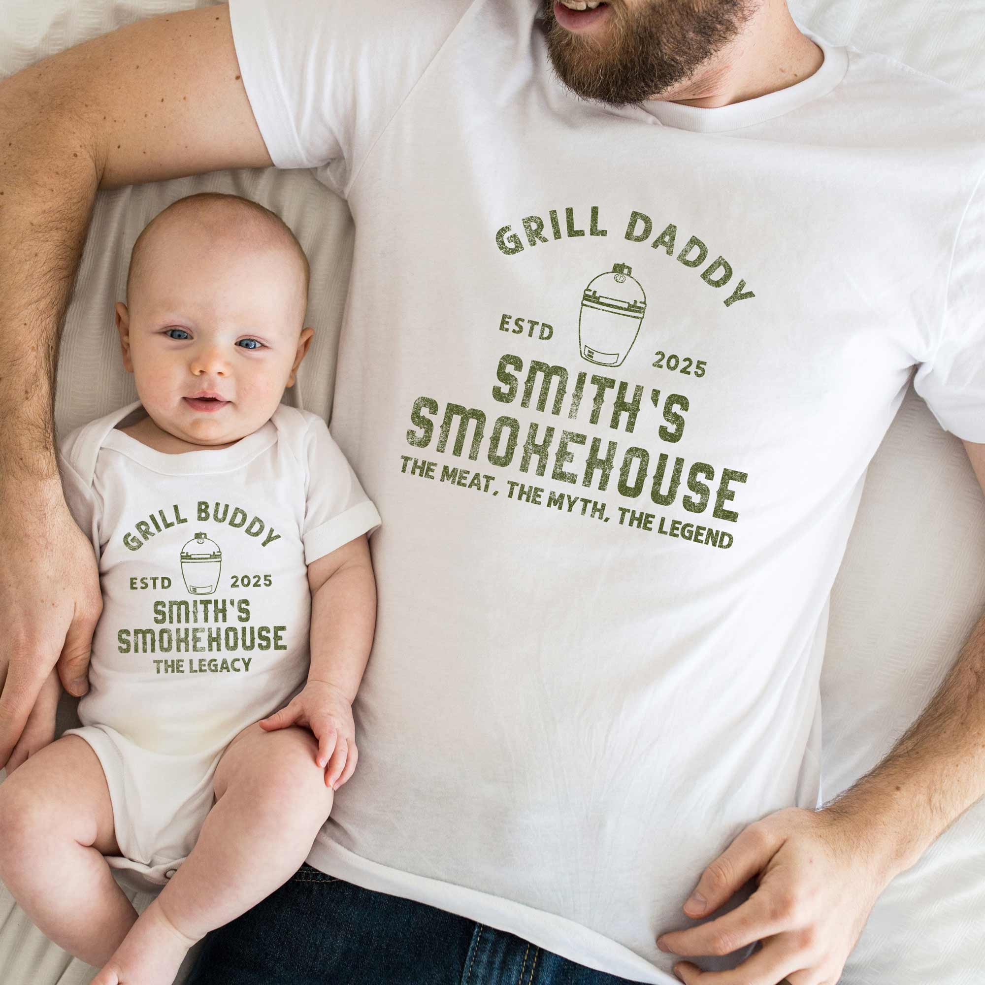 a father and baby laying on a bed, wearing matching dad and son shirts. A grill buddy onesie: meat smoker in white, and a grill daddy shirt in white