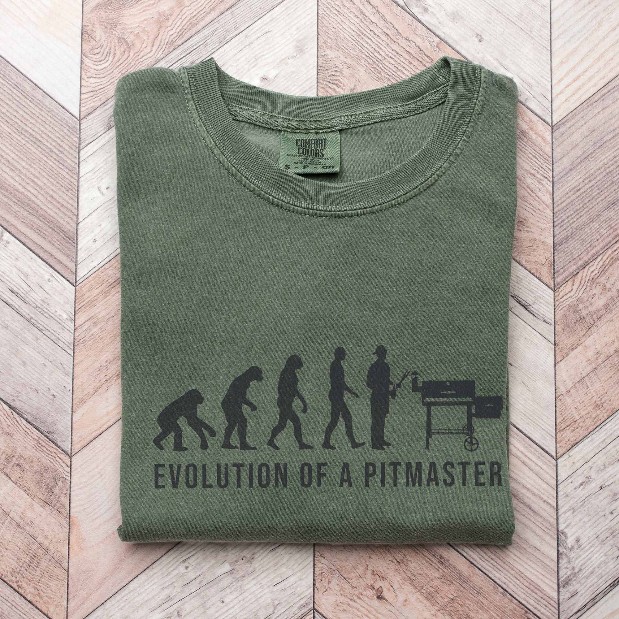 an Evolution Of A Pitmaster Shirt in moss, folded up on a wood background
