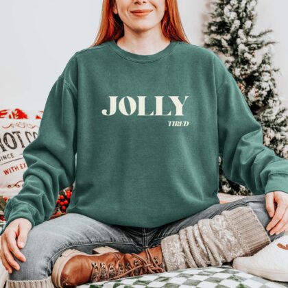 a young woman sitting on a bed in a Christmas scene, wearing a Jolly Tired Sweatshirt in Blue Spruce