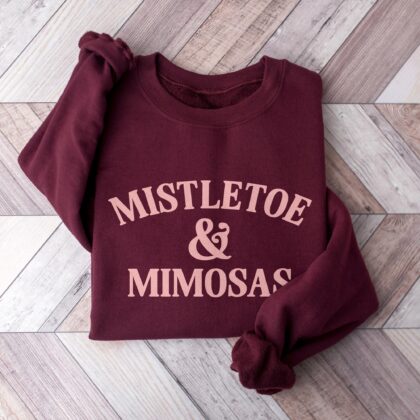 a Mistletoe And Mimosas Sweatshirt in maroon, folded, sitting on a wood floor