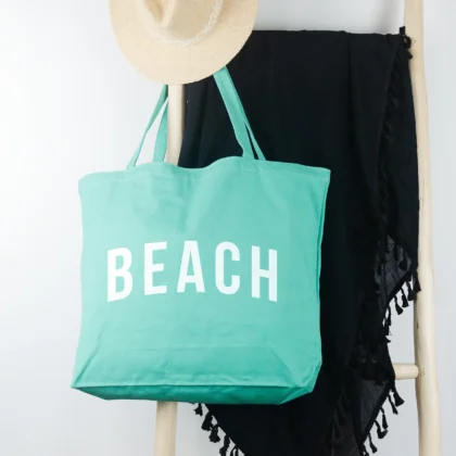 a Pigment beach tote in sea glass, hanging on a ladder, with a sun hat and sarong