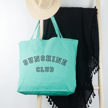 a a Sunshine Club Pigment Tote in sea glass, hanging on a ladder with a sun hat and sarong.