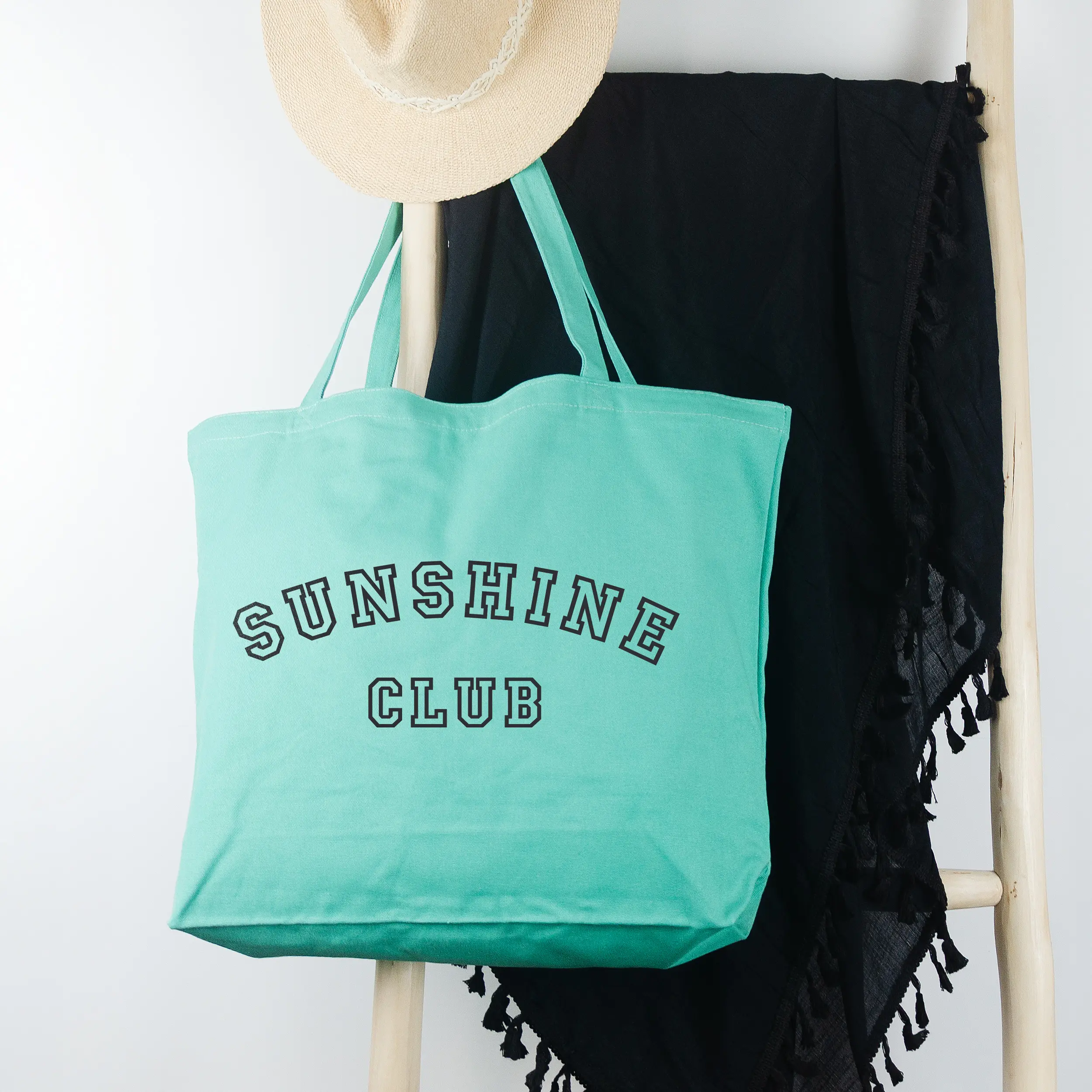 a a Sunshine Club Pigment Tote in sea glass, hanging on a ladder with a sun hat and sarong.